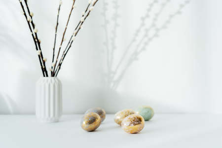 Home interior with easter decor. Pussy willow branches in a vase, golden eggs in different patterns on the white table with light and shadow background. Minimalism, scandinavian style.の写真素材