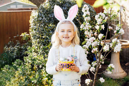 Adorable little girl wearing bunny ears holding a basket with Easter eggs outdoors on spring day....