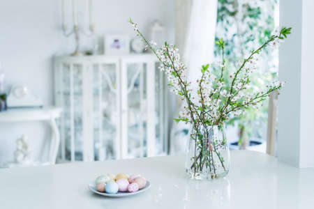 Blooming tree branches in the vase and colored easter eggs on the plate on white kitchen table with classic style interior background. Easter celebration at home. Festive spring composition.の写真素材