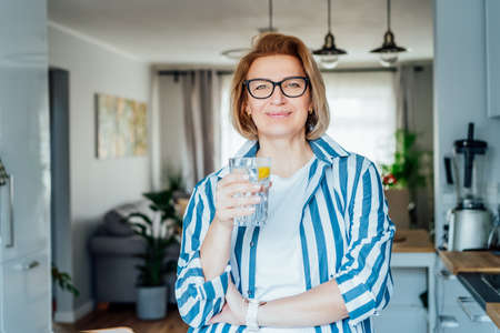 Healthy habit to drink water. Smiling middle age woman with glass of pure water with lemon standing on her kitchen. Control body hydration, Track water balance. Healthy living. Selectivbe focus.の写真素材