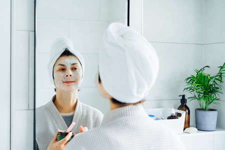 Woman in bathrobe with a towel on her head looking in the mirror and applying facial cosmetic clay...