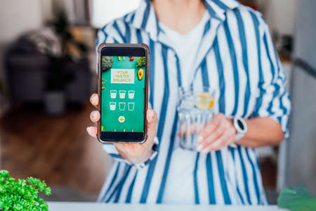 Healthy habit to drink water. Blurred woman with glass of pure water with lemon holding phone with active mobile app to control body hydration, Tracking water balance. Selective focus.の写真素材