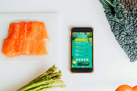 Keto meal diet planning. Top view Smartphone with keto menu in mobile app on table with fresh ingredients, fresh vegetables and salmon fish. Ready to cook. Healthy lifestyle, weight loss. Recipe box.の写真素材
