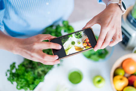 Blogger creating content for social media. Woman making photo, video with just made glass of detox green smoothie and its ingredients on the kitchen table. Healthy dieting, cooking, weight loss blog.の写真素材