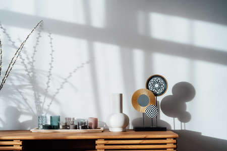 View of a modern minimalist Scandinavian style interior. Candles, ceramic vase and artistic home decor stand on a wooden console under sunlight and shadows on a white gray wall. Living room design.の写真素材