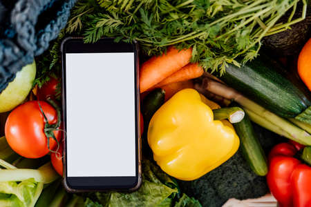 Top view closeup fresh vegetables and phone with blank white screen on white background. Mockup for online mobile application for diet program, food delivery, weight loss. Recipe box. Selective focus.の写真素材