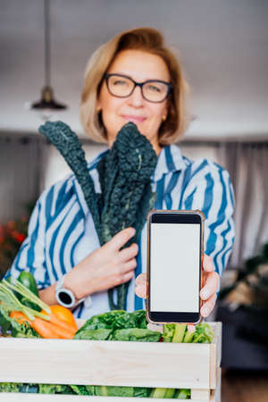 Woman holding phone with blank white screen and fresh kale above wooden box with vegetables. Mockup with copy space for food delivery, diet program, weight loss, recipe box mobile app. Vertical cardの写真素材