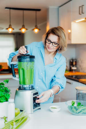 Middle aged woman with blender and green vegetables making detox shake or green smoothie at home. Healthy dieting, eating, cooking. Natural anti aging, weight loss program. Vegan diet. Vertical cardの写真素材