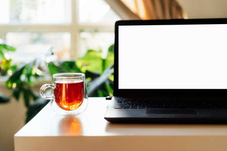 Modern workplace - desk with laptop mockup white screen, cup of tea at work space in home office...