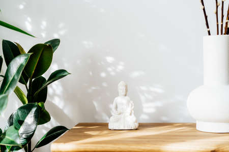 White Buddha statuette home decoration and clay vase standing on wooden cabinet. Minimalist design with green plants and white wall with shadows and highlights. Interior design decor. Selective focus.の写真素材