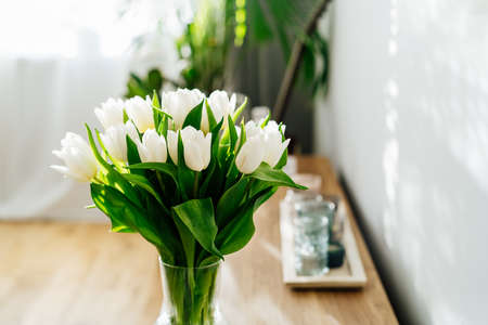 Spring bouquet of tender white tulip flowers in vase stand on a wooden cabinet near light grey wall...