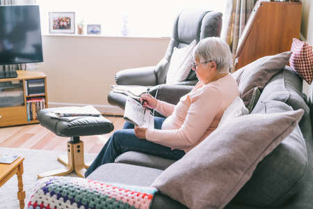 Old senior woman, 80s adult, grandmother, making crossword puzzles in her living room sitting on the sofa. Retirement hobby for training memory. Alzheimers preventive practice. Selective focusの写真素材