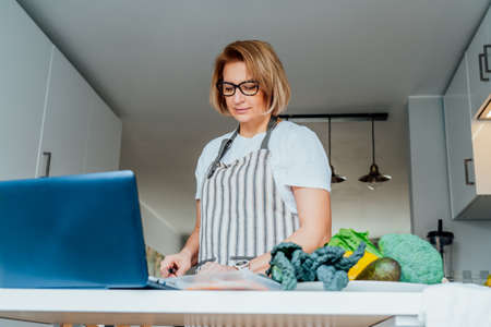 Middle age woman following a cooking tutorial video course on laptop while preparing meal in a kitchen. Woman cooking healthy dish, fish and vegetables on the table. Online recipe. Selective focusの写真素材