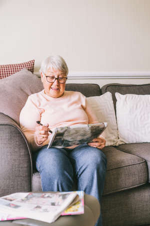 Old senior woman, 80s adult, grandmother, making crossword puzzles in her living room sitting on the sofa. Retirement hobby for training memory. Alzheimers preventive practice. Selective focusの写真素材