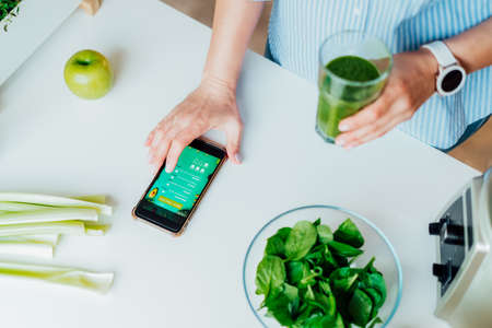 Close up woman with green smoothie fills, making notes in the meal treaker on her phone on the kitchen table. Healthy diet and balanced eating, weight loss online app program. Selective focusの写真素材