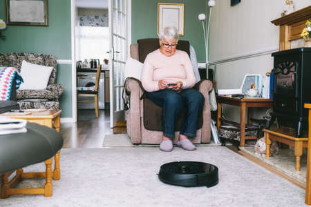 Senior woman using a robot vacuum cleaner through phone settings while sitting on armchair at home. Mature older lady holding mobile phone and control cleaning. Smart home. Seniors and technologyの写真素材