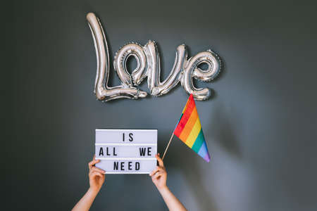 LGBTQ pride month background. Male hands holding lightbox with Text Love is all we need under silver foil balloon with rainbow flag on gray wall background. Film effect tonning. Selective focusの写真素材