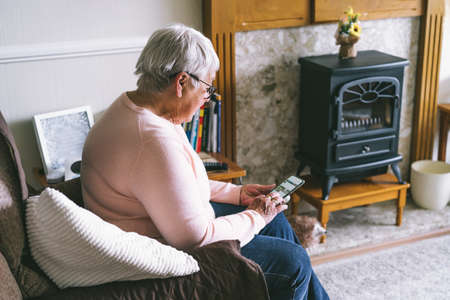 Senior woman using smartphone sitting on armchair at home. Relaxed mature older lady holding mobile...
