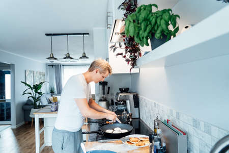 A young man is cooking cheese pancakes in a modern light kitchen at home. Man preparing pancakes for frying on the pan. Housework and home duties. Culinary hobby. Selective focusの写真素材