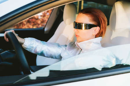 Fashion cyberpunk girl drive a supercar. Red hair woman with futuristic eyeglasses sits by the car...