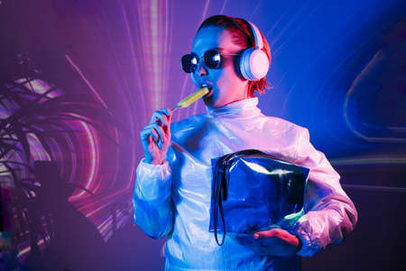 Portrait of a cyberpunk girl in sunglasses and headphones in neon light. Futuristic woman licking a lollipop in holographic clothes on the background of waves Concept. fashion, futuristic generation.の写真素材