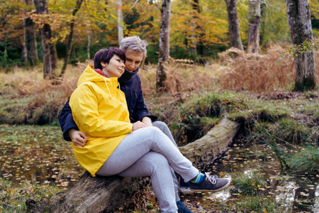 Couple in love sitting on a fallen tree near river, looking at the forest and enjoying the moment. Feeling harmony, reunion with nature in autumn. Relax, personal fulfillment. Romantic time together.の写真素材