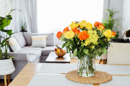 Kitchen counter table with focus on vase with yellow and orange roses on it with blurred background of modern cozy living room with couch and green plants. Open space home interior design. Copy spaceの写真素材