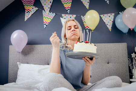 Sad, upset woman in pajama and party cap thinking eat or not to eat birthday cake sitting on the bed in decorated bedroom. Celebrates birthday alone concept. Selective focusの写真素材