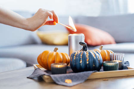 Female hand lighting a candle with a match in autumn cozy composition for hygge home mood. Pumpkins and candles on the tray with gray napkin on the coffee table in the living room. Selective focus.の写真素材