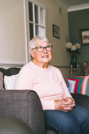 Portrait of smiling senior, mature retired grandmother in eyeglasses, elderly woman smiling sitting on sofa at home enjoying retirement. Vertical card. Selective focusの写真素材
