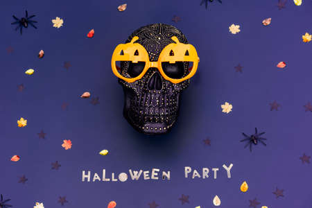 Black decorated human skull wears funny pumpkin glasses and Halloween party lettering made of golden letters on the dark purple blue background with autumn leaves sequins and spiders. Selective focus.の写真素材