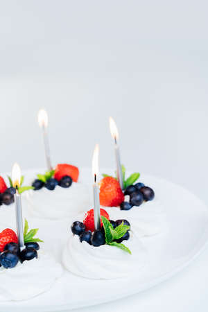 Meringue dessert Pavlova cake nests with fresh strawberries and blueberries with mint on the white plate with burning candles. Happy Birthday cake idea. Vertical card. Copy space. Selective focusの写真素材