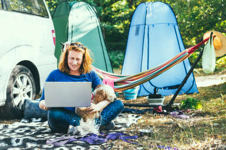 Adult woman use laptop computer sitting with her cockapoo puppy near camper van recreational vehicle with background of tents and nature park. Concept of travel and remote worker people.の写真素材