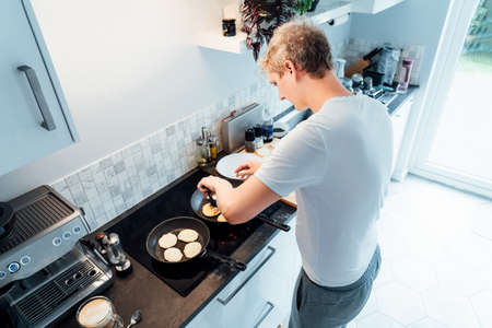 A young man is cooking cheese pancakes in a modern light kitchen at home. Man preparing pancakes for...