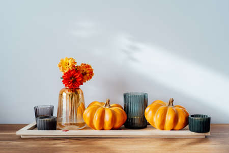 Autumn, fall cozy composition. Orange pumpkins, candles, dahlia flowers in vase on the wooden tray on the table with white wall background. Scandinavian minimalist hygge home decor. Selective focusの写真素材