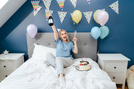 Happy emotional woman in pajama and party cap sitting on the bed with festive cake and champagne bottle in decorated bedroom. Time for yourself Celebration at home. Happy birthday concept.の写真素材