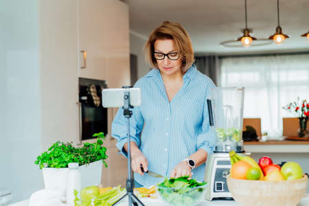 Middle age women is blogging for her wellness lifestyle channel about healthy balanced living in the kitchen of her home. Creating video content for social media with phone camera on a tripodの写真素材