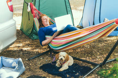 Adult woman working on laptop pc while lying in hammock, with her cockapoo puppy near motorhome on camping trip. Female living on camper car with animals and travel the world. Remote worker peopleの写真素材