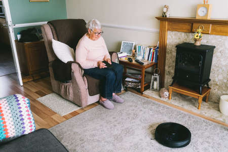 Senior woman using a robot vacuum cleaner through phone settings while sitting on armchair at home. Mature older lady holding mobile phone and control cleaning. Smart home. Seniors and technologyの写真素材
