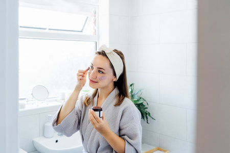 Young woman in bathrobe looking in the mirror and applying facial natural cosmetic clay mask on her face in bathroom. Cosmetic procedures for skin care at home. Beauty self-care. Selective focusの写真素材