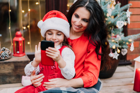 Smiling mother with a daughter in Santa hat congratulate grandparents via video chat while sitting on house porch with decorated Christmas tree. Family time. Festive mood. Selective focus.の写真素材