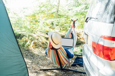 Back view adult woman working on laptop pc with blank screen while lying in hammock near motorhome on camping trip. Female living on camper car and travel the world. Remote worker people.の写真素材