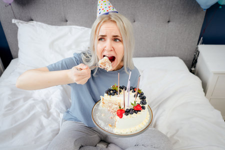 Sad, upset woman in pajama and party cap eating by spoon her birthday cake sitting on the bed in...