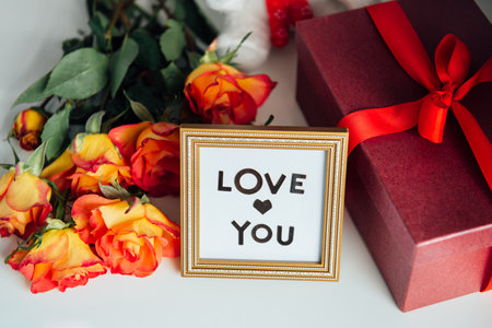 Close up frame with Love you message, gift box with red ribbon and a bouquet of roses flowers on the bedside table. Valentines day, birthday, womens day surprise. Selective focus.の写真素材