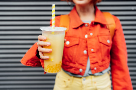 Glass of fruity sugar flavored tapioca pearl bubble tea with straw in hand of no face hipster fashion woman in bright clothes on the gray striped wall background. Selective focus. Copy spaceの写真素材