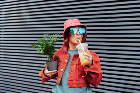 Hipster fashion women in bright clothes, heart shaped glasses, bucket hat drinking fruity sugar flavored tapioca pearl bubble tea and holding green potted plant on the gray striped wall backgroundの写真素材
