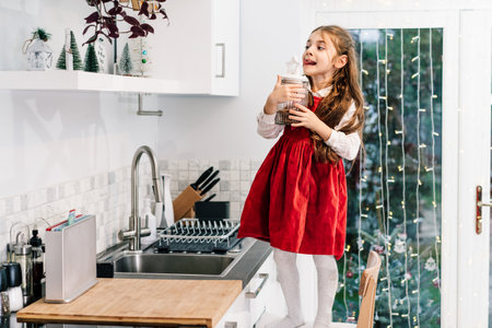 Happy smiling cute little girl kid standing on the chair and reached jar with chocolate festive cookies on the shelf on decorated for winter Christmas holidays modern kitchen. Overeating sweetsの写真素材