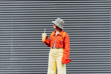 Stylish girl in bright clothes drinking sugar flavored tapioca bubble tea while walking near gray striped urban wall. Full length portrait of fashionable hipster girl. Street fashion. Copy spaceの写真素材