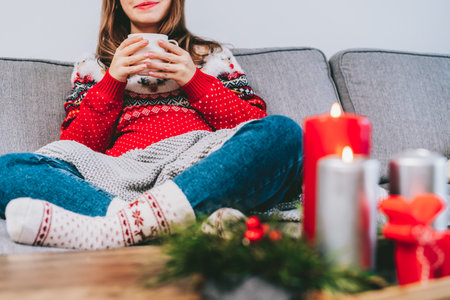 Cropped woman in ugly sweater and warm woolen socks on sofa holding cup of hot drink with cozy Christmas mood candles decor for hygge home on the coffee table. home coziness. Winter and Xmas holidaysの写真素材