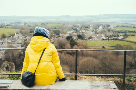 Back view woman in warm bright clothes sitting on bench on the view point and looking down at the medieval English town. Cold season travel. Relax and personal fulfillment. Copy space.の写真素材